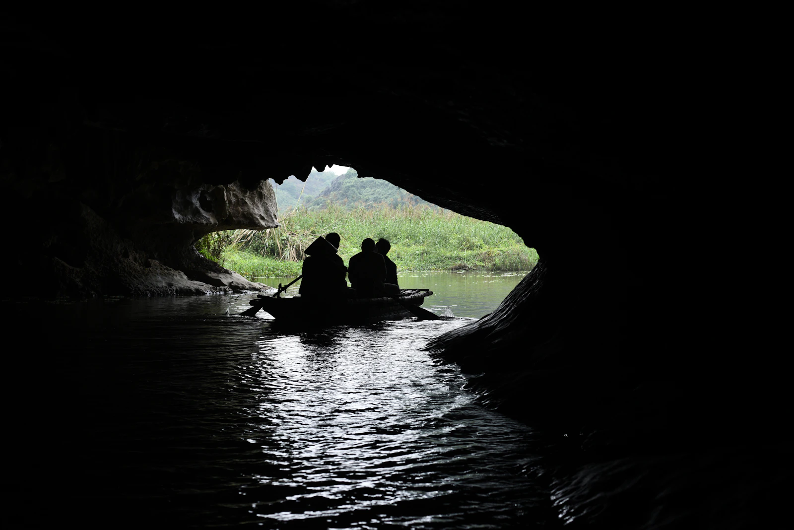 Ruderboot als Silhouette vor einem Höhlenausgang im Van-Long-Naturreservat; draußen Wasserpflanzen und Kalkberge